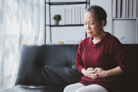 Senior woman sitting in the living room at home and showing body aches, aging sickness, body aches of elderly people, organ degeneration.の写真素材