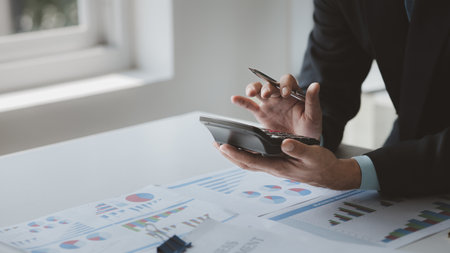 A businessman is using a calculator to calculate financial numbers, he is sitting in his private office, the businessman examines the financial data from the corporate finance chart.の写真素材