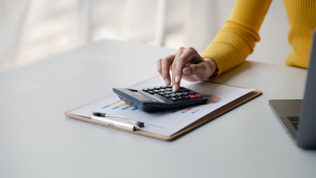 Businessman using a calculator to calculate numbers on a company's financial documents, she is...