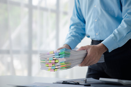 A person in an office with a huge stack of papers, he is a company employee, managing paperwork and...