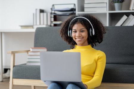 American woman sits in living room on weekends, she is wearing headphones listening to music and...