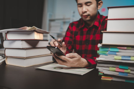 Asian male businessman working in the room. Startup company employee is pressing smartphone to view information, he is in office at night, working overtime, lots of work, hard work. Overtime concept.の写真素材
