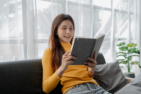 Asian woman in the living room and reading a book, female company employee on vacation, using...