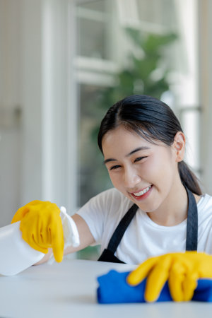 Asian female cleaner wiping down tables with cleaning spray, Wear rubber gloves and an apron and work with a happy smile, Use a towel to wipe the table, cleaning idea.の写真素材