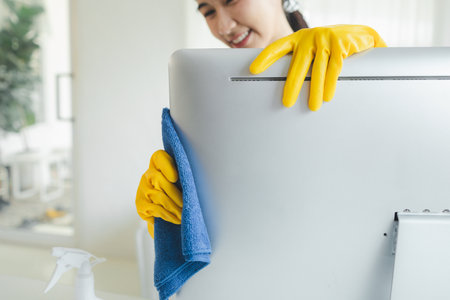Asian female cleaner, Wear rubber gloves and an apron and work with a happy smile, Cleaning staff wiping down office equipment, Wipe the monitor clean with a towel and sanitizer, cleaning idea.の写真素材