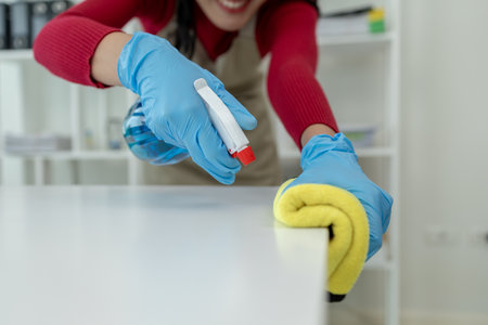 Asian female cleaner wiping down tables with cleaning spray, Wear rubber gloves and an apron and work with a happy smile, Use a towel to wipe the table, cleaning idea.の写真素材