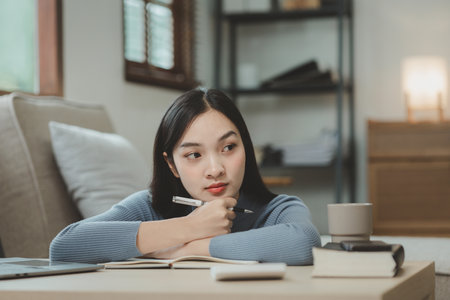 A student is sitting in the living room doing homework, Sitting and looking for information and making reports to send to teachers in the house, Recording various information In the book at apartment.の写真素材