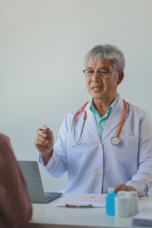 male medical practitioner reassuring a patient, Doctor and patient in conversation, Asian male doctor consulting patient, Professional physician wearing white coat talking to mature womanの写真素材
