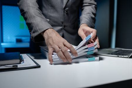 An employee held a large pile of documents in his hand, Large piles of work documents on desks are being sorted by employees, Important documents are stacked.の写真素材