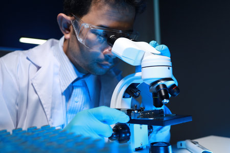 A talented scientist sits in his lab while using a microscope to look for microscopic organisms in a...