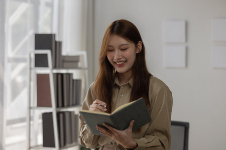 an Asian businesswoman is writing detail from the last meeting, there is a beautiful employee is smiling in the workplace, taking a note into the book witch is the first draft of reportの写真素材