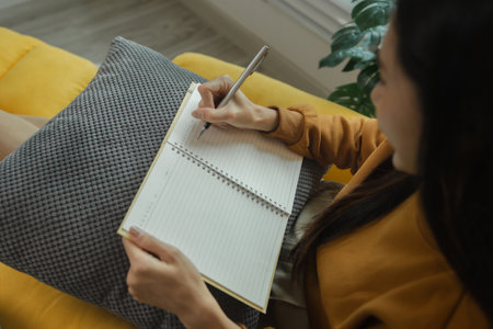 An Asian woman is sitting on the sofa in the living room. She is keeping a diary to record past memories and experiences. Keeping a diary is a leisure activity that allows us to reflect on things.の写真素材