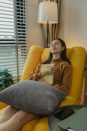 An Asian woman is sitting on the sofa in the living room. She is keeping a diary to record past...