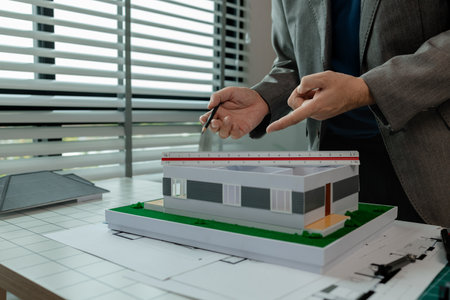 An architect works in the office, an engineer is using a tool to take measurements of a house model so that it matches the blueprint, there is a house model on the architect's deskの写真素材