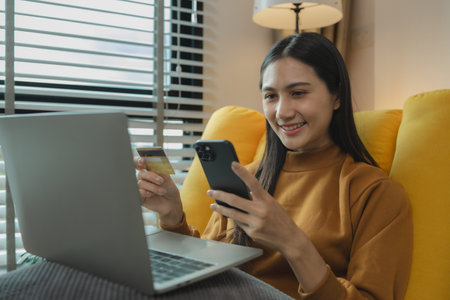 Asian woman sitting on the sofa in the living room, she is shopping online by paying by credit card, shopping on an online shopping platform, paying by credit card or debit card online.の写真素材