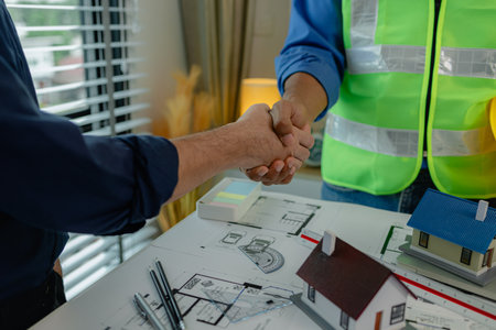 engineer and architect are shaking hands to celebrate because they just finished big construction project, two engineers do handshake after the meeting, blueprint is on the deskの写真素材