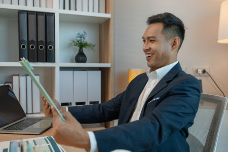 middle aged professional businessman wearing white shirt and suit happily smiling in office, a middle-aged, dark-haired businessman in a suit is happily working at his desk in the officeの写真素材