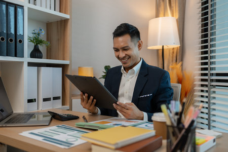 middle aged professional businessman wearing white shirt and suit happily smiling in office, a middle-aged, dark-haired businessman in a suit is happily working at his desk in the officeの写真素材