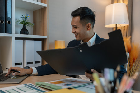 middle aged professional businessman wearing white shirt and suit happily smiling in office, a middle-aged, dark-haired businessman in a suit is happily working at his desk in the officeの写真素材
