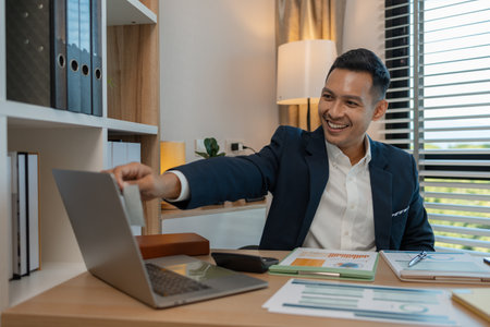 middle aged professional businessman wearing white shirt and suit happily smiling in office, a middle-aged, dark-haired businessman in a suit is happily working at his desk in the officeの写真素材