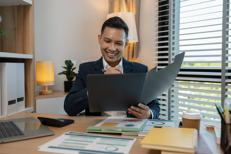 middle aged professional businessman wearing white shirt and suit happily smiling in office, a middle-aged, dark-haired businessman in a suit is happily working at his desk in the officeの写真素材