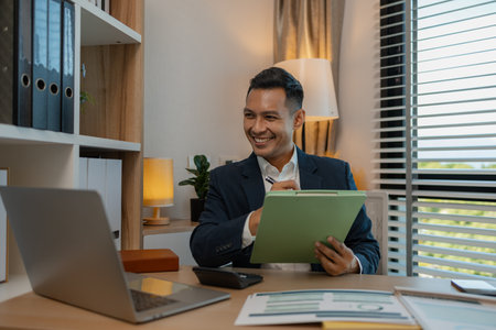middle aged professional businessman wearing white shirt and suit happily smiling in office, a middle-aged, dark-haired businessman in a suit is happily working at his desk in the officeの写真素材