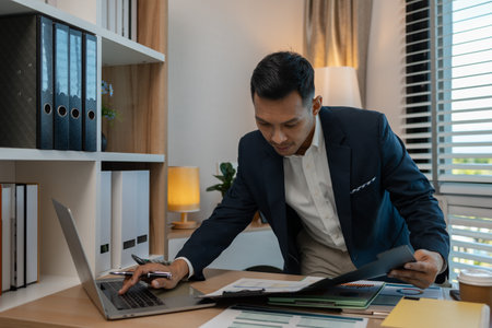 middle aged professional businessman wearing white shirt and suit happily smiling in office, a middle-aged, dark-haired businessman in a suit is happily working at his desk in the officeの写真素材