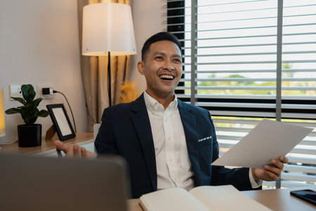 middle aged professional businessman wearing white shirt and suit happily smiling in office, a middle-aged, dark-haired businessman in a suit is happily working at his desk in the officeの写真素材