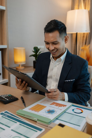 middle aged professional businessman wearing white shirt and suit happily smiling in office, a middle-aged, dark-haired businessman in a suit is happily working at his desk in the officeの写真素材
