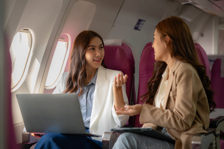 two beautiful asian female friends are traveling abroad for vacation by plane and while sitting on the plane they use electronic devices to entertain themselves during the journeyの写真素材
