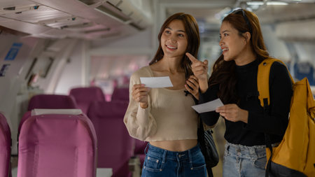 two beautiful female friends are traveling abroad on a vacation by plane, they walk to find their seats and store their bags in the overhead compartment, passenger, airplane, vacationsの写真素材