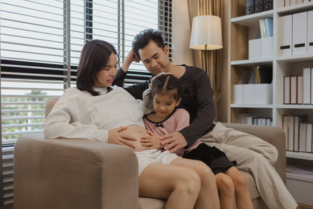 lovely family spend time together in their house, a pregnant mother sits on the couch with her husband and daughter enjoying a happy time together on a weekendの写真素材
