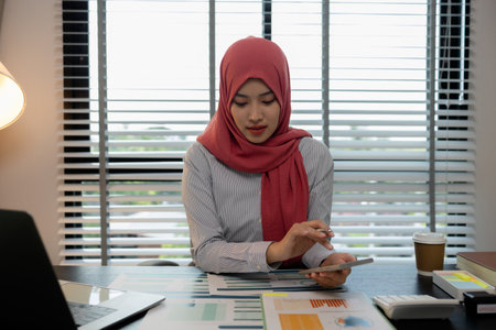 a muslim female employee is working on financial and accounting documents at a company in her private office, hijab, marketing, the female muslim company president is sitting in her office workingの写真素材