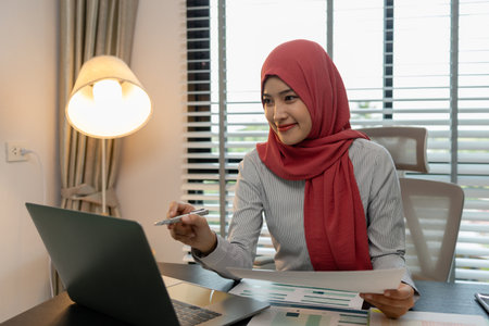 a muslim female employee is working on financial and accounting documents at a company in her private office, hijab, marketing, the female muslim company president is sitting in her office workingの写真素材