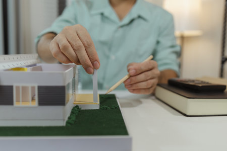 An architect in a light green shirt working on a modern house model at a desk, carefully assembling...