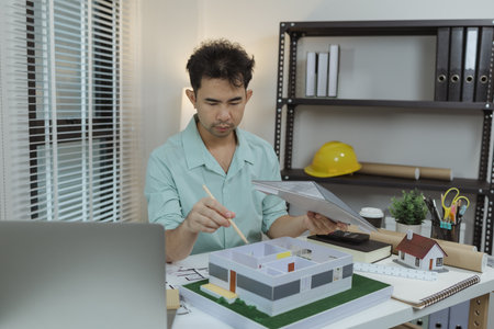 Asian male architect is analyzing a modern house model while holding the roof structure and pointing...