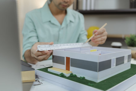 Asian male architect sitting at a desk, measuring a modern house model with triangular ruler and...