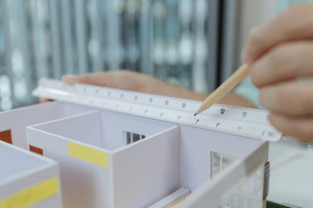 Asian male architect sitting at a desk, measuring a modern house model with triangular ruler and pencil. Office setup includes design tools, blueprints, and safety gear, suggesting architectural work.の写真素材