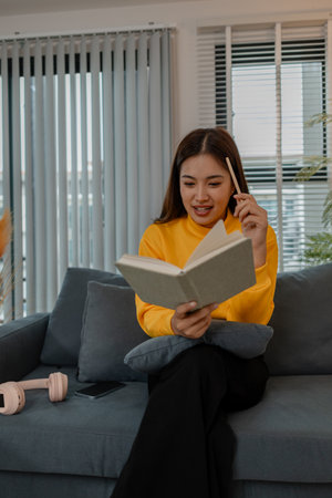 Asian woman in yellow sweater reading a book while sitting on a gray sofa at home. She looks focused...