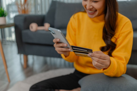 Asian woman holding a smartphone and credit card while making an online purchase at home. She sits...