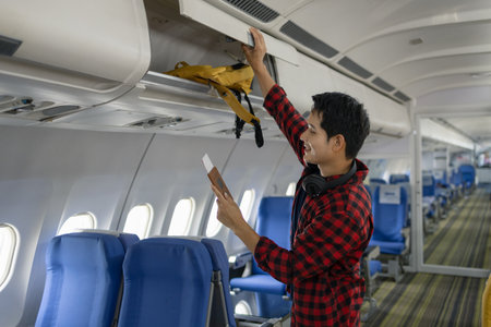 Young Asian man placing a yellow backpack into an overhead compartment on an airplane. He looks...