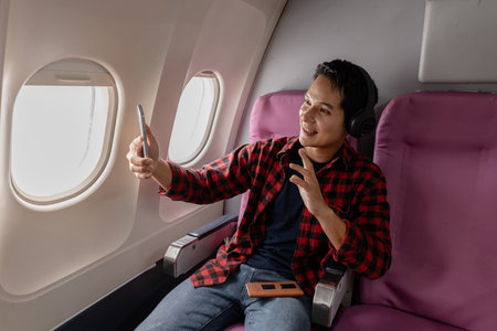 A cheerful young man enjoys his flight while using his smartphone and wearing headphones. Sitting by...