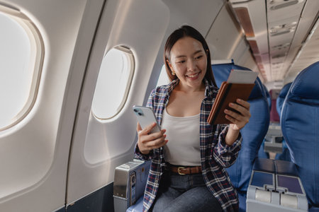 A young woman sitting in an airplane seat, smiling while holding her phone and passport, taking selfies and enjoying the flight. She looks relaxed and excited for her journey ahead.の写真素材