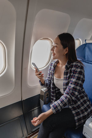 Young Asian woman using a smartphone while sitting in a window seat on an airplane. She looks cheerful and engaged with her phone, possibly texting, watching something, or using social media.の写真素材