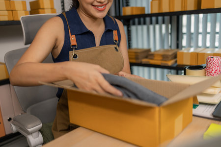 Smiling young female entrepreneur packing clothes into a parcel box, happily preparing merchandise...
