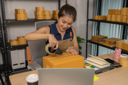 Young female entrepreneur carefully sealing package with tape, preparing merchandise orders, managing shipment and delivery for her successful online clothing business at her home workspace.の写真素材