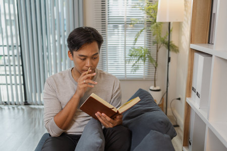 A young man is relaxing on a sofa, reading a book with a calm expression. He enjoys quiet time and...