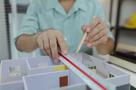 Asian male architect sitting at a desk, measuring a modern house model with triangular ruler and pencil. Office setup includes design tools, blueprints, and safety gear, suggesting architectural work.の写真素材