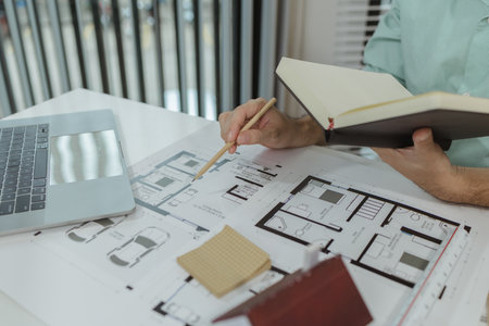 Architect reviewing house floor plan while holding a notebook and pencil. The workspace includes a...