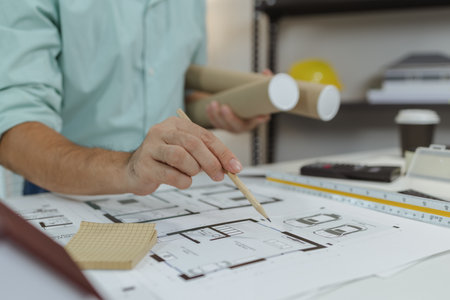 An architect pointing at house floor plans with a pencil while holding blueprint rolls. The desk is filled with architectural tools, scale ruler, and model materials during planning.の写真素材